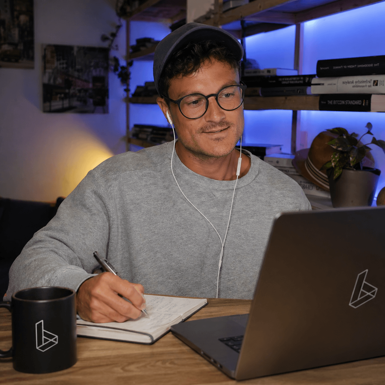 Alec am Schreibtisch mit Laptop, Notizblock und Tasse, Regal mit u. a. The Bitcoin Standard im Hintergrund