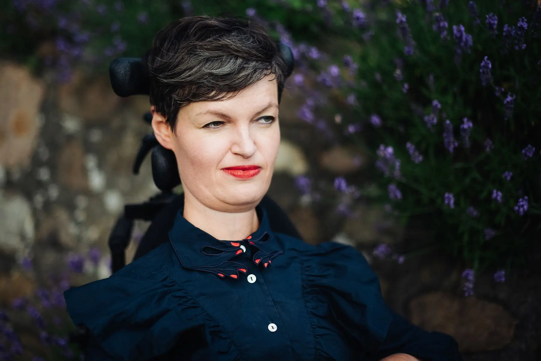 Porträtfoto einer Frau mit kurzem dunklem Haar und rotem Lippenstift, die in einem Rollstuhl sitzt und nachdenklich in die Ferne schaut.